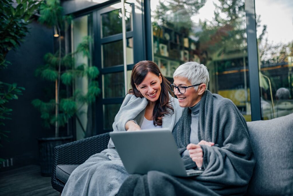 A woman helps an elderly woman on her laptop outside