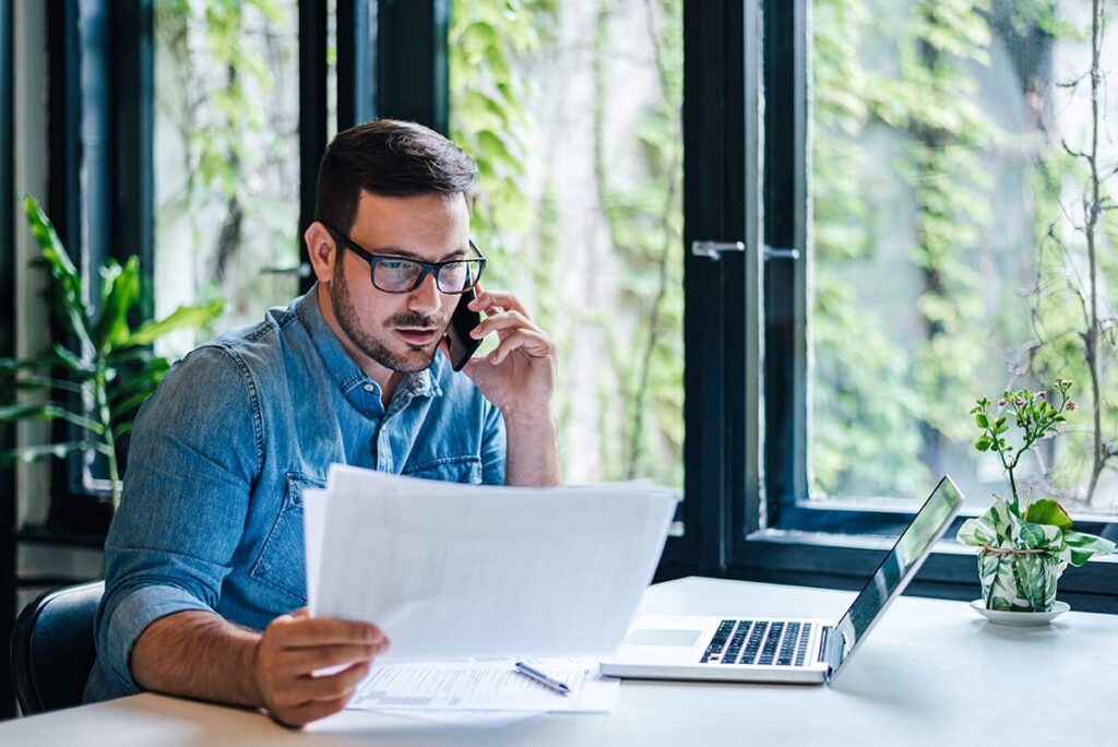 A man on a phone call while looking at his laptop and looking through papers